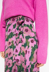 Suéter de punto rosa combinado con una falda de talle alto que presenta grandes patrones florales rosas sobre un fondo verde oscuro, atada en la cintura.