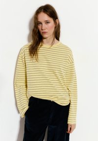 Camicia a maniche lunghe gialla a righe con linee grigie, dalla texture morbida. Abbinata a pantaloni di velluto a coste scuri. Design minimalista, vestibilità rilassata.