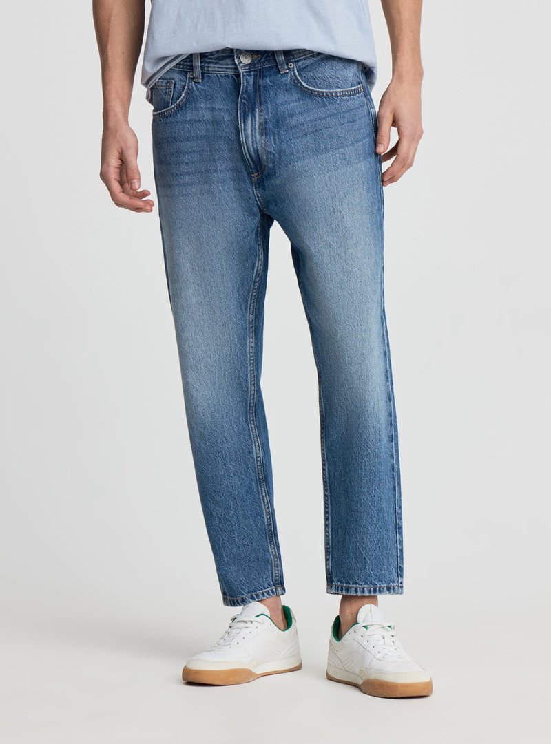 Uomo con jeans azzurri chiari e sneakers bianche in piedi su uno sfondo semplice.