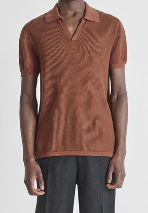 Homme portant un polo en maille marron à manches courtes avec un motif texturé et un pantalon gris foncé sur un fond blanc uni.