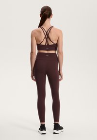 Brun sport-bh med korsade band och matchande leggings, båda i ett slätt, stretchigt material. Svarta träningsskor kompletterar outfiten.