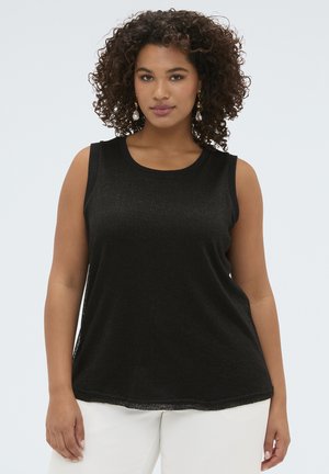 Femme aux cheveux bouclés portant un haut noir sans manches et un pantalon blanc, regardant droit devant sur un fond clair.