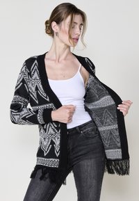 Cardigan in bianco e nero con un motivo a righe, texture lavorata a maglia, davanti aperto e dettagli frangiati all'orlo. Indossato sopra una canottiera bianca.