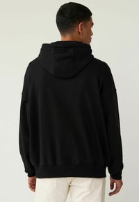 Sort oversized hættetrøje med snoretræk i hætten, ribbede cuffs og kant, samt en glat stoftekstur, set fra ryggen.