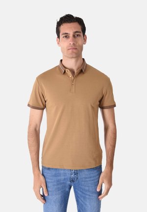 Uomo che indossa una polo beige a maniche corte con rifiniture marroni, jeans blu, in piedi davanti a uno sfondo chiaro e uniforme.