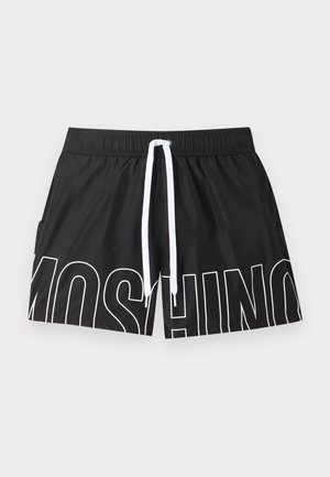 Man zonder hemd met donker haar die zwarte zwemshorts met witte trekkoord en zwarte Moschino slippers draagt op een witte achtergrond.