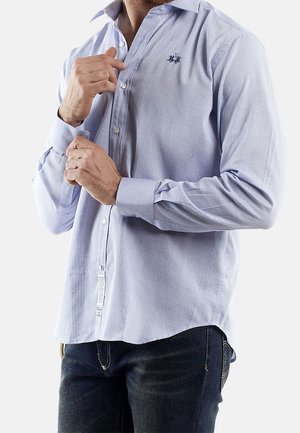 Mężczyzna zapinający mankiety jasnoniebieskiej koszuli z długim rękawem, z delikatnym wzorem w kropki i małym haftowanym logo na piersi, noszący ciemne dżinsy.