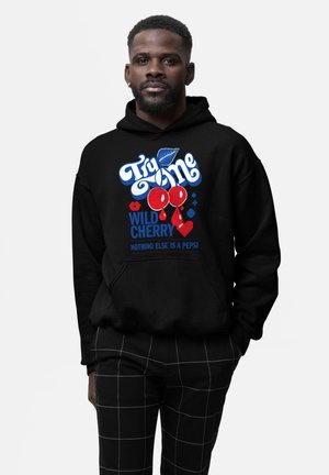 Schwarzer Hoodie mit roten Kirschen und dem Text "Try Me Wild Cherry Nothing Else Is a Pepsi", getragen von einem Mann in schwarz karierter Hose mit den Händen in den Taschen.