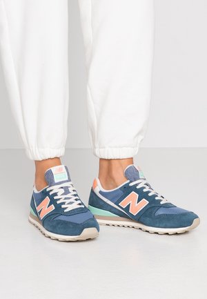 Pies con zapatillas New Balance azules y durazno con cordones blancos, combinados con pantalones deportivos blancos sobre un fondo liso.