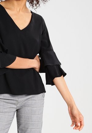 Femme portant un chemisier noir à col en V avec des manches évasées à niveaux et un pantalon gris à carreaux, posant avec un bras croisé sur sa taille.