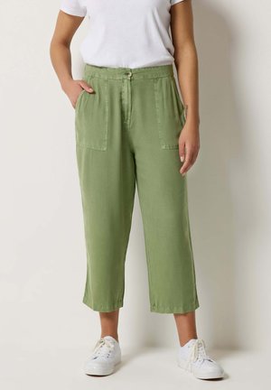 Person trägt olivgrüne, verkürzte Hosen mit großen Fronttaschen, weiße Turnschuhe und ein weißes, kurzärmeliges Hemd, das in die Hose gesteckt ist, und steht aufrecht.