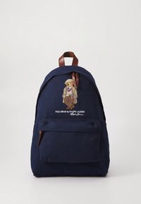 Granatowy płócienny plecak z elementami z skóry, z haftowanym misiem w garniturze na froncie i kieszenią na zamek poniżej.