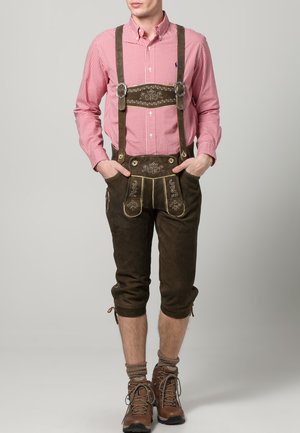 Mann in traditionellen braunen Lederhosen mit floralen Stickereien, rotem karierter Hemd und braunen Wanderschuhen.