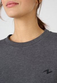 Grijze geribbelde sweatshirt met een ronde halslijn en een klein zwart logo aan de linkerkant. Gladde textuur met subtiele details.