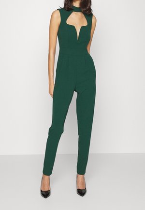 Zielony kombinezon z elastycznego materiału, z wysokim kołnierzem, wycięciami na klatce piersiowej i zwężanymi nogawkami. Noszony z czarnymi szpilkami.