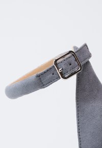Bride de chaussure en daim gris avec une boucle carrée argentée et plusieurs trous de réglage, présentée sur un fond clair uni.