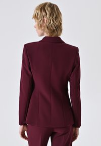 Femme avec des cheveux blonds courts et ondulés, portant un blazer ajusté bordeaux foncé et un pantalon assorti, de dos contre un fond clair uni.