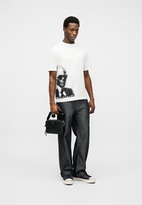 Νεαρός άνδρας με λευκό μπλουζάκι με γραφικά, μαύρο τζιν παντελόνι φαρδύ, μαύρα αθλητικά παπούτσια, κρατώντας μια μικρή μαύρη τσάντα σε απλό λευκό φόντο.