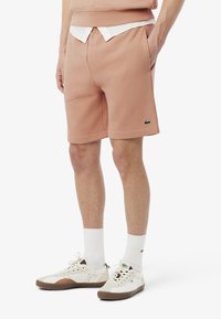 Pantalones cortos de algodón rosa claro con un ajuste relajado, bolsillos frontales y un logo verde de Lacoste. Combinados con calcetines blancos y zapatillas deportivas retro.