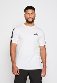 Camiseta de manga corta blanca hecha de algodón. Presenta el logotipo negro de Puma en el pecho y acentos negros con un patrón en los hombros.