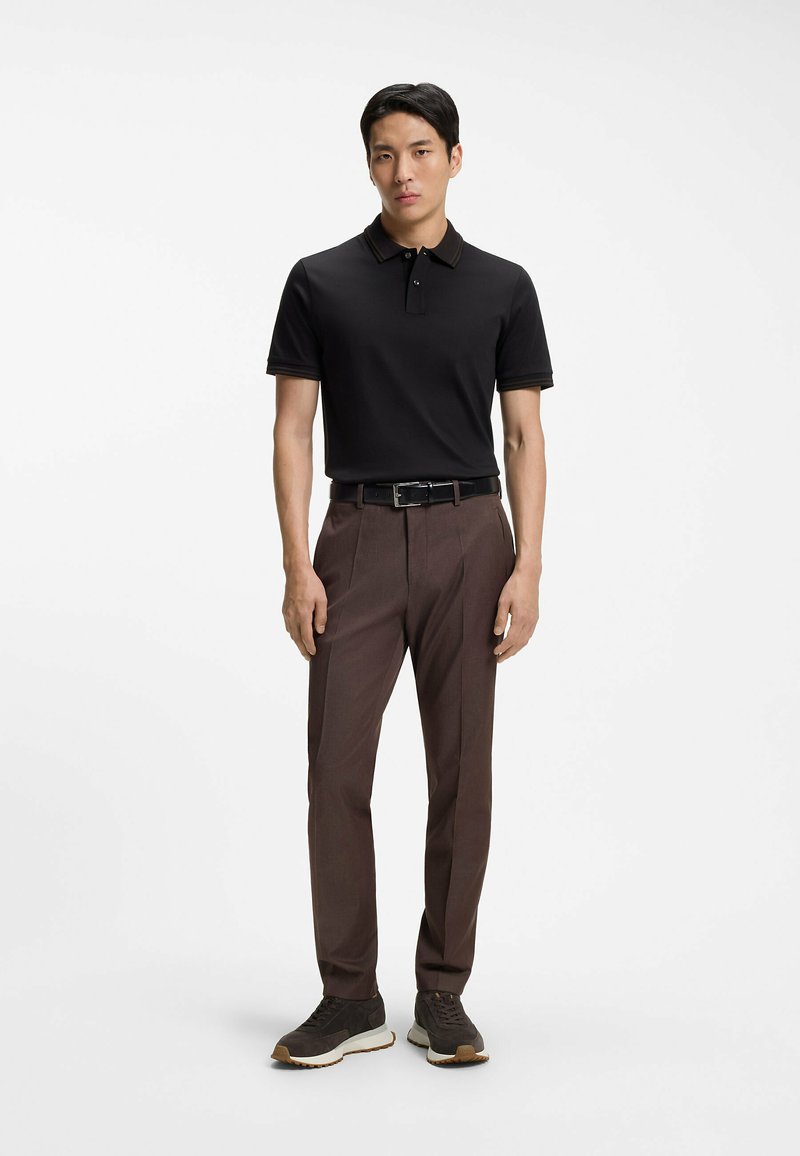 Jeune homme debout, portant un polo noir, un pantalon marron sur mesure avec une ceinture noire, et des baskets foncées sur un fond blanc.