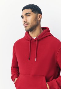 Rode katoenen hoodie met een voorzak, verstelbare trekkoorden en geribbelde boorden. De stof lijkt zacht en glad, met een casual ontwerp.