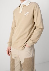 Beige långärmad polo med vit krage och broderad sköldlogotyp. Slät tyg med ribbade manschetter, tillsammans med ljusbeiga kordbyxor med fickor.