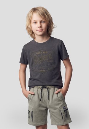 Niño rubio con las manos en los bolsillos, vistiendo una camiseta gráfica oscura y pantalones cortos cargo claros, de pie contra un fondo gris liso.