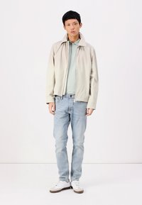 Light beige jacket over a light blue polo, paired with faded blue jeans and white sneakers. Smooth fabric, relaxed fit, minimal design.