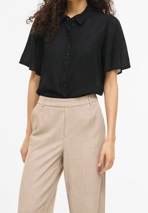 Femme portant un chemisier noir à manches courtes avec des boutons, rentré dans un pantalon beige taille haute à jambes larges.