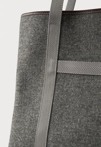 Κοντινό πλάνο μιας γκρι τσάντας από τσόχα με υφή γκρι διάτρητους χειρολαβές και καφέ δερμάτινη επένδυση κατά μήκος της πάνω άκρης.