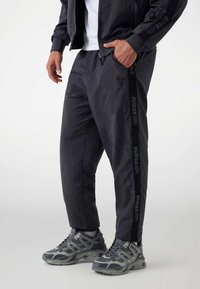 Mann in schwarzen Jogginghosen und passender Jacke mit "GUESS"-Logo an der Seite der Hose, kombiniert mit grauen Sportschuhen.