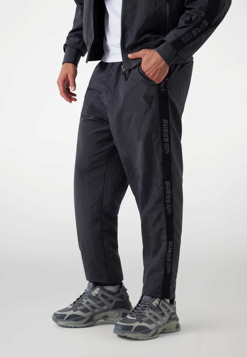 Mann in schwarzen Jogginghosen und passender Jacke mit "GUESS"-Logo an der Seite der Hose, kombiniert mit grauen Sportschuhen.