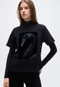 Νεαρή γυναίκα που φοράει μαύρο μπλουζάκι με μεγάλο γραφικό μαύρο κύκνο, από πάνω από μακριά μαύρα μανίκια παγιέτας, στέκεται μπροστά σε απλό φωτεινό φόντο.