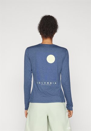 Personne aux cheveux bouclés portant une chemise bleue à manches longues avec un graphisme rond de soleil et le texte "Columbia Sportswear" au dos.