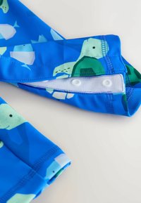 Costume da bagno blu con una stampa acquatica giocosa con pesci e tartarughe. Presenta una texture liscia e un dettaglio di chiusura a scatto bianca.