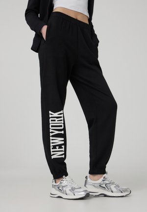 Pantaloni da jogging neri con vita elastica e tasche laterali, con la scritta bianca "NEW YORK" stampata verticalmente sulla gamba sinistra.