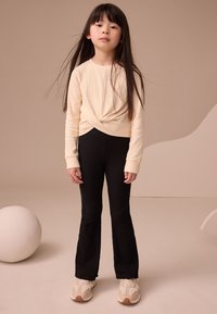 Haut à manches longues beige côtelé avec un design noué, associé à un pantalon évasé côtelé noir et des baskets beige avec des accents clairs.