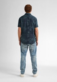 Camisa de manga corta en color navy con un patrón de hojas tropicales verdes, combinada con jeans azul claro y zapatillas blancas, vista desde la parte de atrás.