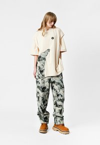 Mujer vestida con una camiseta beige de gran tamaño con un estampado abstracto, pantalones sueltos a juego y zapatos gruesos en tono beige, de pie contra un fondo blanco.