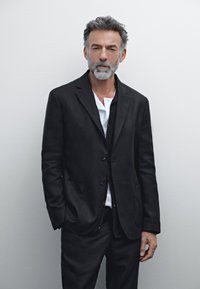 Schwarzer Leinenanzug mit einem taillierten Blazer und passenden Hosen. Der Blazer hat ein einreihiges Design, zwei Taschen und ein weißes Hemd darunter.