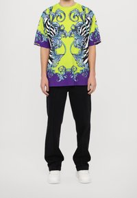 Tričko s krátkým rukávem s odvážným žlutým a fialovým designem s propracovanými vzory, pruhy zebry a uvolněným střihem. Černé kalhoty a bílé tenisky.