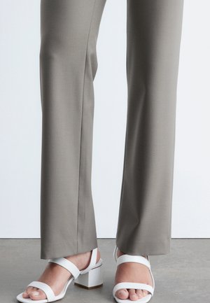 Jambes portant un pantalon droit gris clair et des sandales blanches à talons blocs ouverts à l'avant, debout sur un sol gris.