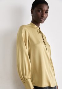 Blusa a maniche lunghe di un delicato giallo chiaro con un collo alto, dettaglio annodato al colletto e maniche arricciate. Tessuto morbido e fluido.