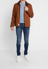 Brun bomullsjacka med en mörk krage, bärs över en ljusblå tröja och blå smala jeans, komplett med vita sneakers.