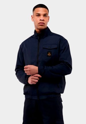 Jonge man die een donkerblauw jasje met rits en een borstzak met logo-patch draagt en zijn linkermouw aanpast.