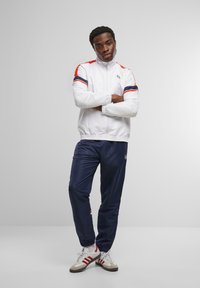 White track jacket with red and navy accents, paired with navy track pants. Both have elastic cuffs, showcasing a sporty, active wear design.