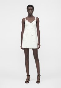 Donna che indossa un mini abito bianco senza maniche con applicazioni floreali e sandali neri con tacco alto, in piedi su uno sfondo bianco.