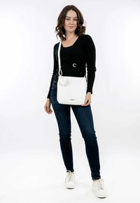 Sac bandoulière en cuir blanc avec une finition lisse, de forme rectangulaire, et une étiquette en forme de cœur. Modèle portant un haut noir et un jean bleu.