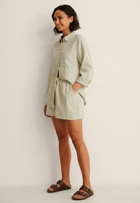 Light olive green linen shirt with long sleeves and a chest pocket, paired with matching shorts. Brown sandals complete the look.